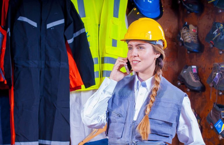 Geberb Reinigung Frau mit Helm telefoniert, im Hintergrund Schutzkleidung und Sicherheitsausrüstung.